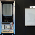 左の「BS-DU」（信号処理・制御など）と「BS-RRH」（無線部）が基地局設備で、従量はそれぞれ約20kg程度。端末は現在FPGAボードで実現しており、右のキャビネットの中に収められている