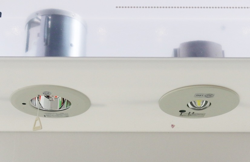 ハロゲン電球非常用照明器具「IEM-13221HN」（左）とLED非常用照明器具「LEDEM13221」（右）。「IEM-13221HN」に付いているヒモは、定期点検時に重りを吊るし、きちんと点灯するかを確認するためのもので、「LEDEM13221」はボタンのワンタッチ操作で点検が可能（撮影：防犯システム取材班）